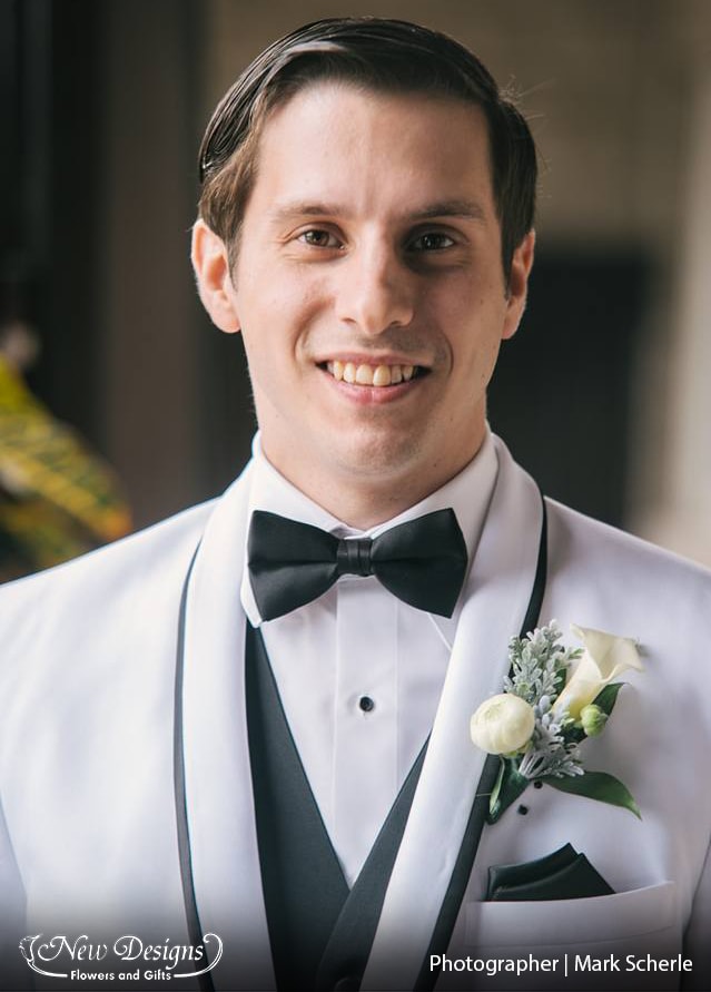 A groom in a crisp white suit, sports a boutonniere with A white calla lilly, and white ranunculus